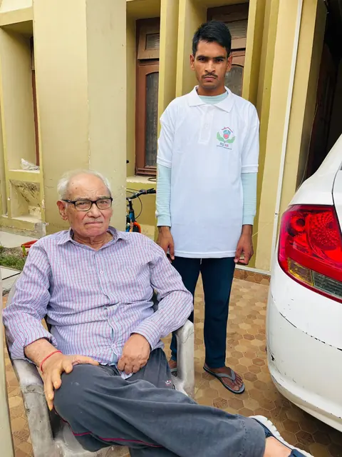 Caregiver standing beside a smiling elderly man seated in a home courtyard