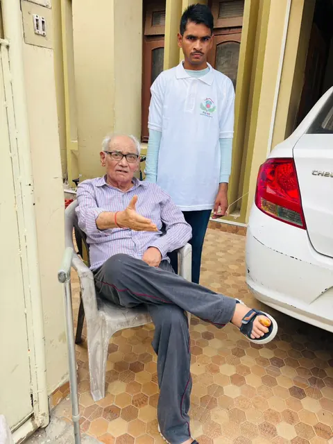 Elderly man seated with a walking stick while a caregiver stands behind him