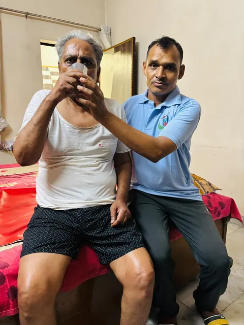 Caregiver helping an elderly man take a drink while sitting on a bed
